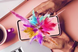 Enhance Your Psychedelic Trip. A top-view of a rose pink table with the corner of a MacBook along the bottom. On the left is the edge of a metallic peach mug with black coffee in it. A person's hands from the right are holding a phone facing up towards the camera. Exploding from the phone screen is a rainbow of what appears to be colorful powder. 
