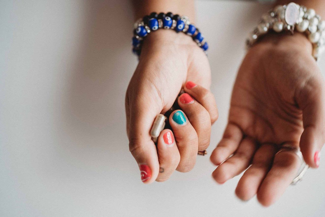 Spiritual Bypassing. Top-down view of hands above a white surface. The left hand on the right of the image, which has an out of focus wide bracelet on the wrist (pearl and silver) is open, facing up. The right hand in the middle of the image which has beaded bracelets on the wrist (one with black beads, and one with blue evil eye beads) is holding psilocybin capsules.