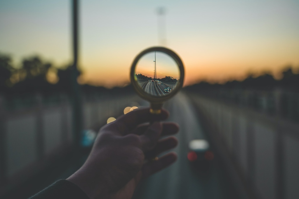 Can Ayahuasca. A person's hand holding a magnifying glass above an out of focus highway, bringing cars in the distance into focus.