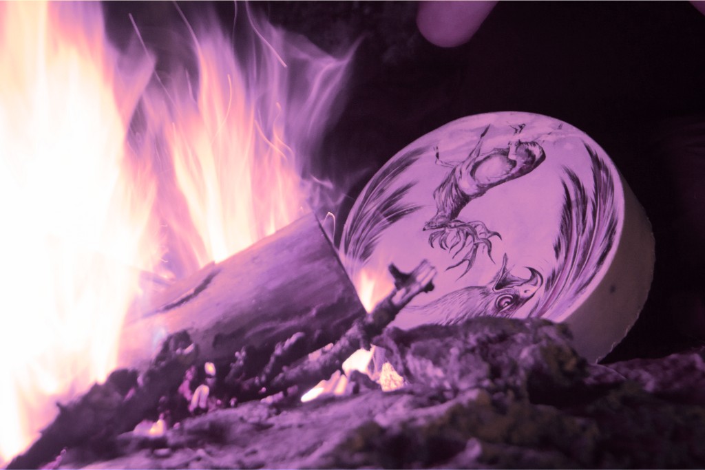The Dark Side of Plant Medicine. Close-up of the edge of an outdoor fire with a shamanic leather tambourine with animal drawings next to it.
