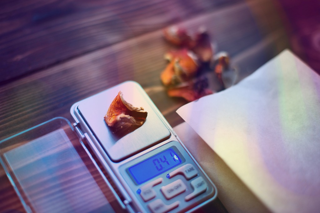 Microdosing for depression. A microgram scale with psilocybin being weighed out on it, with some more of them in the background. There is a piece of paper to the right of the scale, and soft colourful sheer patterns over the whole image. 