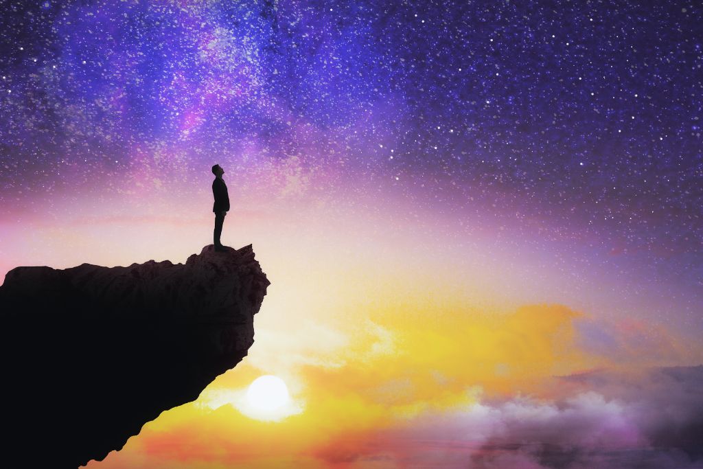 Search for Purpose. Silhouette of a cliff jutting out from the left with man standing on it looking out and up over an expansive background of a starry sky merged with a sunset.