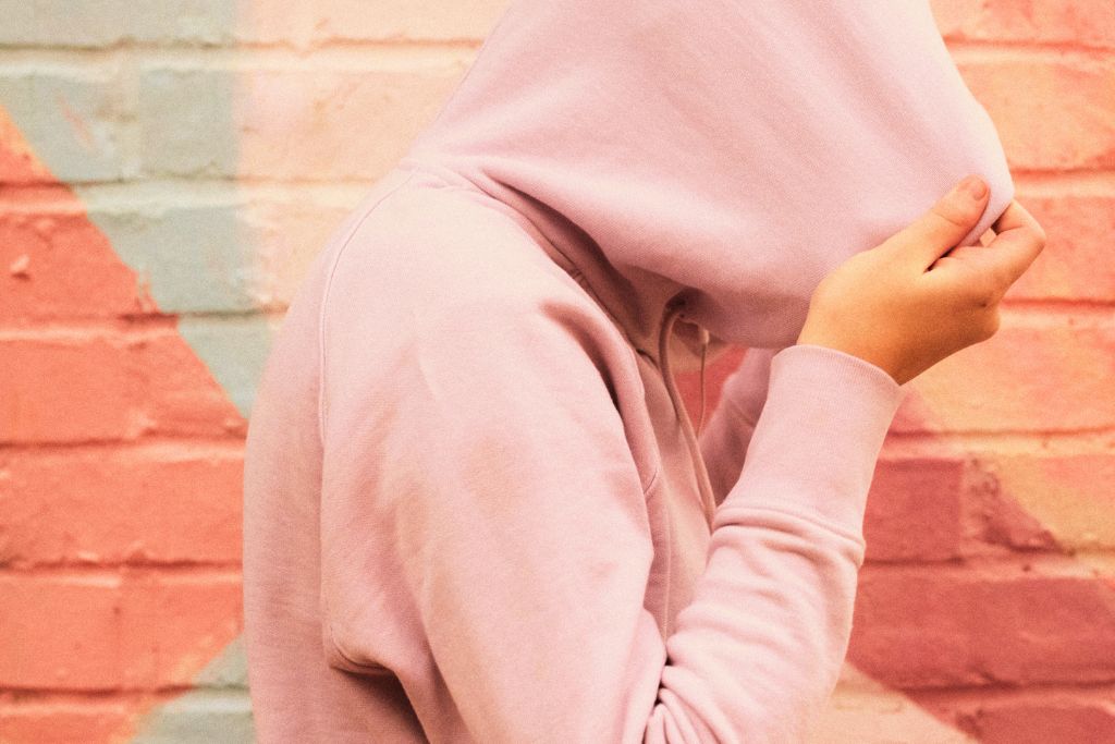 Why Someone Who Looks Healthy May Have An Eating Disorder. Image is a side-view of a person walk to the right side of the image, wearing a pink hoodie with the hood pulled over their head completely so you can only see their hands holding the hood. The wall behind them is painted with pastel colours in bold shapes.