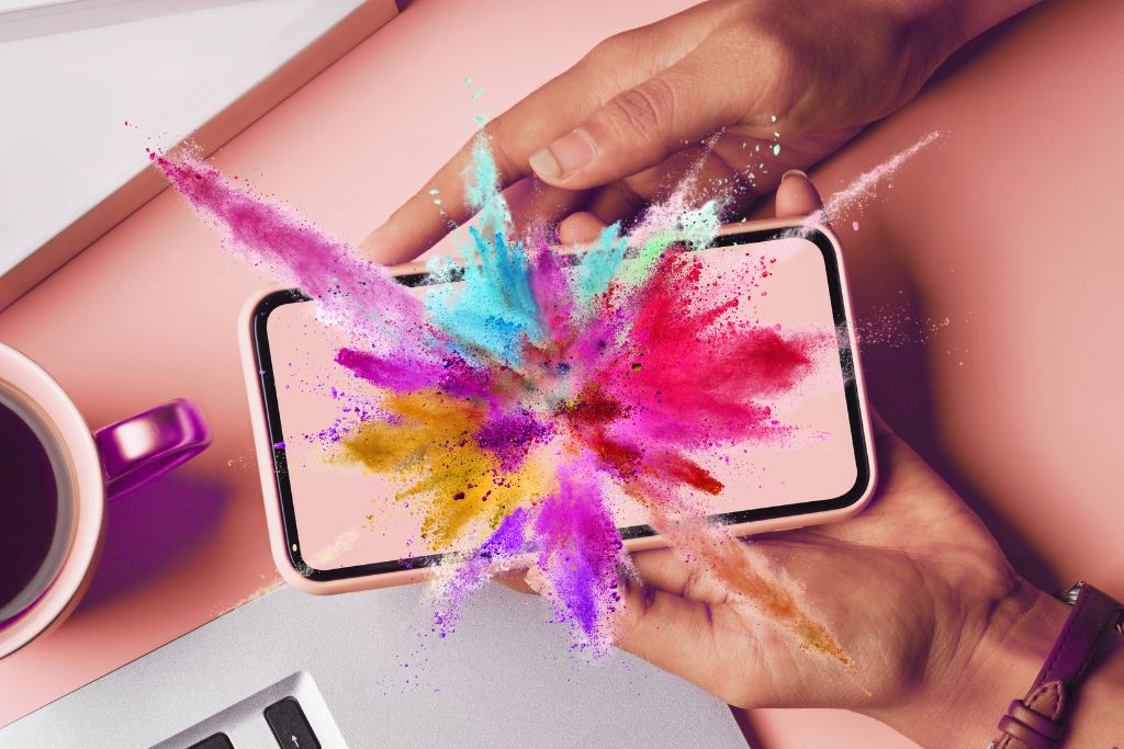 Enhance Your Psychedelic Trip. A top-view of a rose pink table with the corner of a MacBook along the bottom. On the left is the edge of a metallic peach mug with black coffee in it. A person's hands from the right are holding a phone facing up towards the camera. Exploding from the phone screen is a rainbow of what appears to be colorful powder.