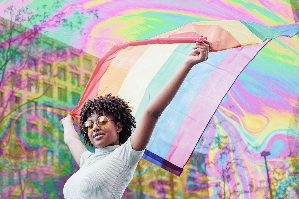 Racialized trauma. An image of a BIPOC person walking down a street holding up a pride flag. They are wearing sunglasses. There is a feint psychedelic rainbow pattern over the image.