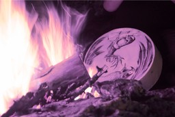 The Dark Side of Plant Medicine. Close-up of the edge of an outdoor fire with a shamanic leather tambourine with animal drawings next to it.