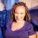 Image of Courtney Watson shows a Black woman with wrapped locks and wearing a purple t-shirt in the sun smiling at the camera