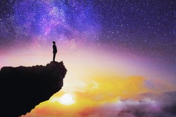 Search for Purpose. Silhouette of a cliff jutting out from the left with man standing on it looking out and up over an expansive background of a starry sky merged with a sunset.