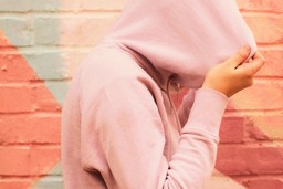 Why Someone Who Looks Healthy May Have An Eating Disorder. Image is a side-view of a person walk to the right side of the image, wearing a pink hoodie with the hood pulled over their head completely so you can only see their hands holding the hood. The wall behind them is painted with pastel colours in bold shapes. 