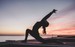 Person doing a yoga pose with on leg bent in a lunge and their arms raised together above their head, sunset in the background. Yoga is a compliment to KAP