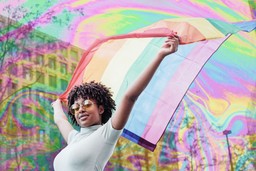Racialized trauma. An image of a BIPOC person walking down a street holding up a pride flag. They are wearing sunglasses. There is a feint psychedelic rainbow pattern over the image.