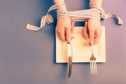 3 Most Common Types of Eating Disorders. Picture is a top-down view of a plate, with a person's hands above the plate, holding a knife and fork. The person's hands are wrapped together with a measuring tape.