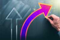 Value shifts. A hand draws a bright, curved neon-pink and orange arrow on a dark chalkboard, rising upward beside a faint, blurred white arrow in the background.