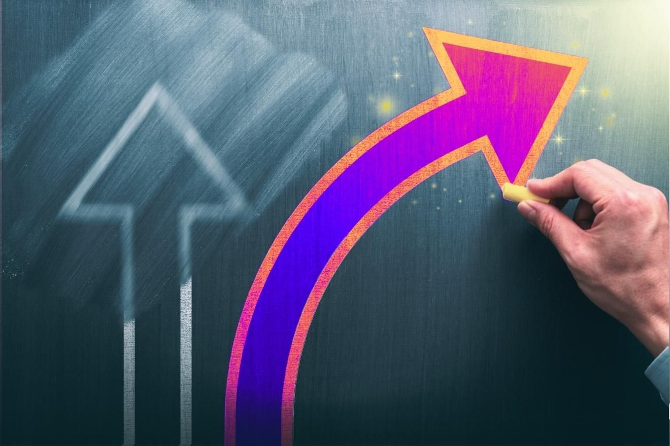 Value shifts. A hand draws a bright, curved neon-pink and orange arrow on a dark chalkboard, rising upward beside a faint, blurred white arrow in the background.