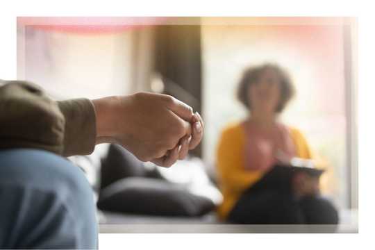 Preparation and integration. A close-up of a person's clasped hands during a therapy session, with a therapist sitting out of focus in the background holding a notebook, symbolizing preparation, integration, and therapeutic support.