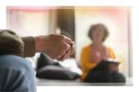 Preparation and integration. A close-up of a person's clasped hands during a therapy session, with a therapist sitting out of focus in the background holding a notebook, symbolizing preparation, integration, and therapeutic support.