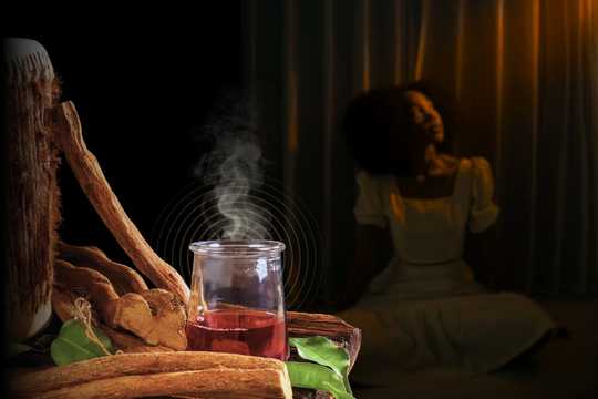 Trauma Healing. A steaming glass jar filled with a crimson herbal ayahuasca concoction, surrounded by dried ayahuasca bark and leaves on a rustic wooden table. In the background, a female-presenting person with an afro hairstyle sits on the floor, draped in soft, warm light, their back to the camera, gazing into the distance.