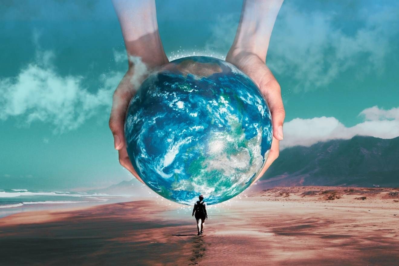 Creative process. A surreal beach scene where a person walks toward a massive Earth held between two giant hands, featuring cool turquoise and sandy tones with soft clouds and mountains in the background.