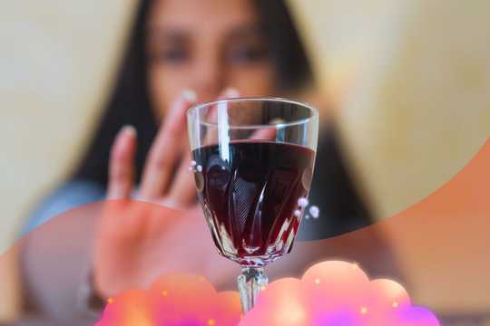 MDMA therapy for alcohol use disorder. The image is of a wine glass with red wine in it. In the background is an out-of-focus person holding up their hand in a "stop" or "no" motion. Around the stem of the wine glass are dreamy pink, peach, and yellow clouds with little sparkles. There is a semi-opaque peach-colored wave-like shape overlayed in front of the photo of the person and behind the wine glass.