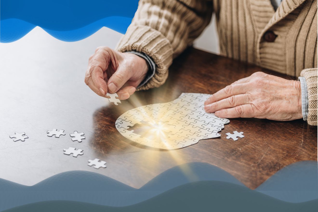 Alzheimer’s disease and related dementias. Image of a person sitting at a table, wearing a tan-colored sweater and building a puzzle of the shape of a human head in profile. There is golden light emanaing from the centre of the puzzle where they are placing the few remaining pieces. There are also blue wave-life shipes along the top of the image, and similar blue-grey wave-like shapes along the bottom.