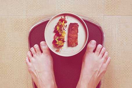 3 Most Common Types of Eating Disorders. Picture is a top-down view of a red scale, showing a person's feet, as well as a plate with food on it on the scale between their feet.