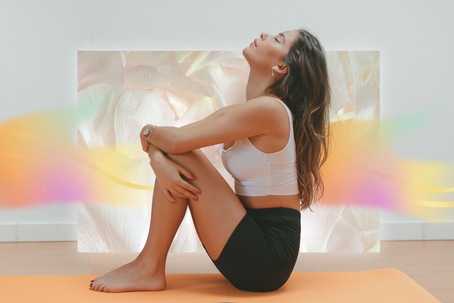 Spiritual Health Professionals. The image features a woman seated on an orange yoga mat in a relaxed, meditative pose. She is wearing a white sports bra and black shorts, with her arms wrapped around her legs and her head tilted back in a calm, upward gaze. The background is a softly lit room with a dreamy, pastel overlay of flowing abstract textures in shades of peach, gold, lavender, and mint green. These ethereal color swirls give the image a spiritual or psychedelic tone, creating a serene and introspective atmosphere. The overall composition emphasizes stillness, inner peace, and gentle transformation.