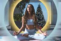 Holiday season self-care. The image features a woman in a meditative pose, sitting cross-legged with her eyes closed, exuding a sense of calm and focus. She is dressed in light blue activewear, with a serene outdoor setting in the background. The composition includes a Christmas tree decorated with ornaments, adding a festive element. A glowing circular halo surrounds her, creating a focal point and imparting a sense of tranquility. The colors are a mix of soft blues, yellows, and greens, enhanced by subtle sparkles, giving the image a harmonious and peaceful atmosphere.