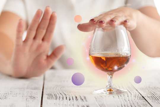 Ketamine therapy for alcohol use disorder. A presumably alcoholic beverage in a whisky glass on an ash-colored wood table. There is a person sitting at the table, only their torso visible, with their left hand, which has red-painted nails covering the glass, and their right hand held up in a "stop" or "no" motion. There is a glowing pink and yellow orb behind the glass, and there are bubble-like circles floating around the glass in gradients of purple, pink, and peach.