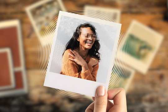 Autobiographical memory. The image shows a hand holding a Polaroid-style photo featuring a woman with dark hair, wearing a warm-toned, textured, orange-brown sweater. Her face is pixelated for anonymity, but her broad smile and crossed arms suggest a joyful, relaxed moment. The background of the Polaroid is a light, neutral wall, while the scene behind the photo is a wooden surface scattered with other blurred photographs, suggesting nostalgia or memory. Warm, earthy tones dominate the color palette, with a soft focus and vignette effect enhancing the intimate, reflective atmosphere of the composition.