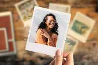 Autobiographical memory. The image shows a hand holding a Polaroid-style photo featuring a woman with dark hair, wearing a warm-toned, textured, orange-brown sweater. Her face is pixelated for anonymity, but her broad smile and crossed arms suggest a joyful, relaxed moment. The background of the Polaroid is a light, neutral wall, while the scene behind the photo is a wooden surface scattered with other blurred photographs, suggesting nostalgia or memory. Warm, earthy tones dominate the color palette, with a soft focus and vignette effect enhancing the intimate, reflective atmosphere of the composition.