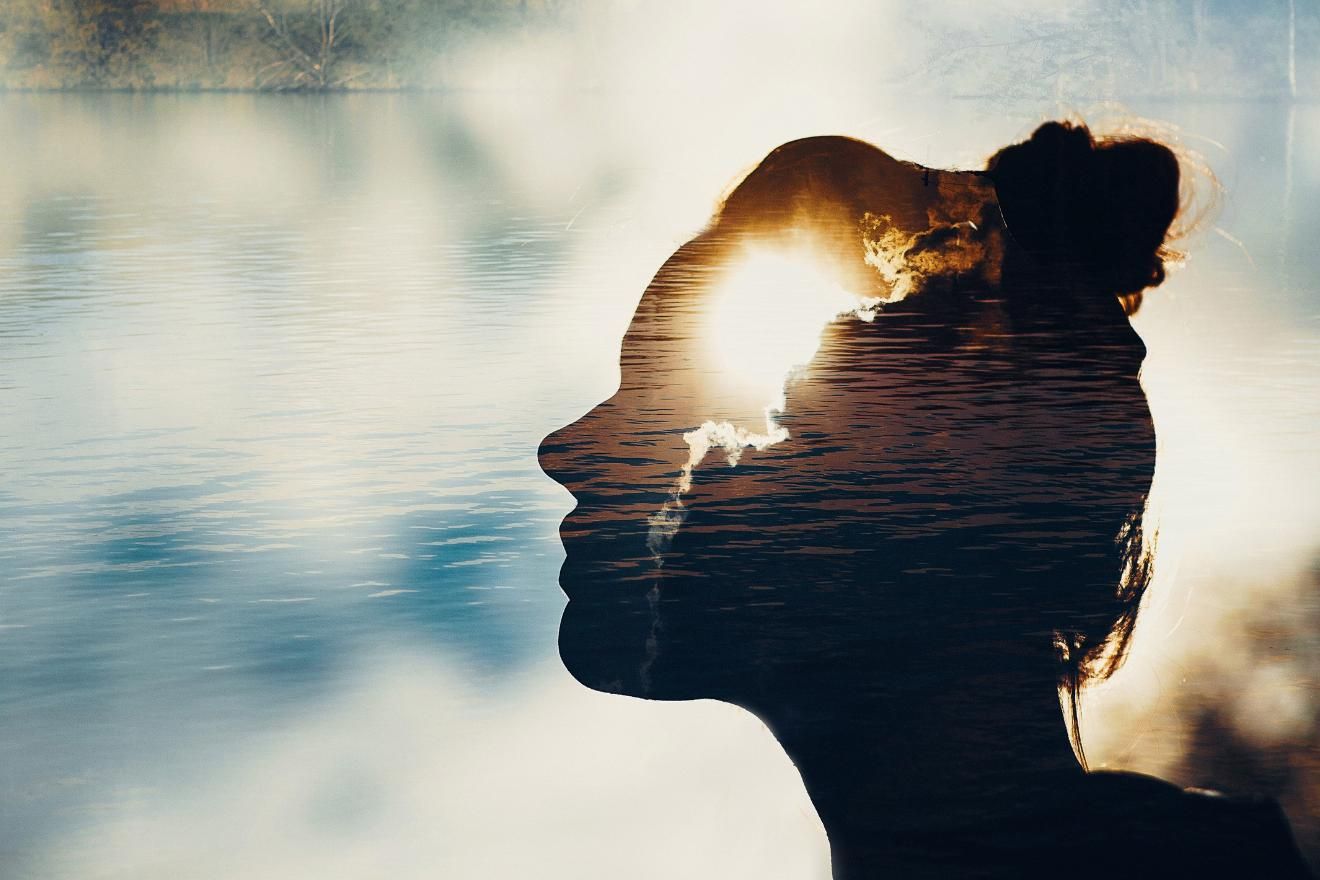 Making Sense of Bad Trips Through Storytelling. Image is a shadow profile of a female presenting person with clouds parting and sun pushing through in the shape near the forehead. The background is of water with a dappled effect over it.