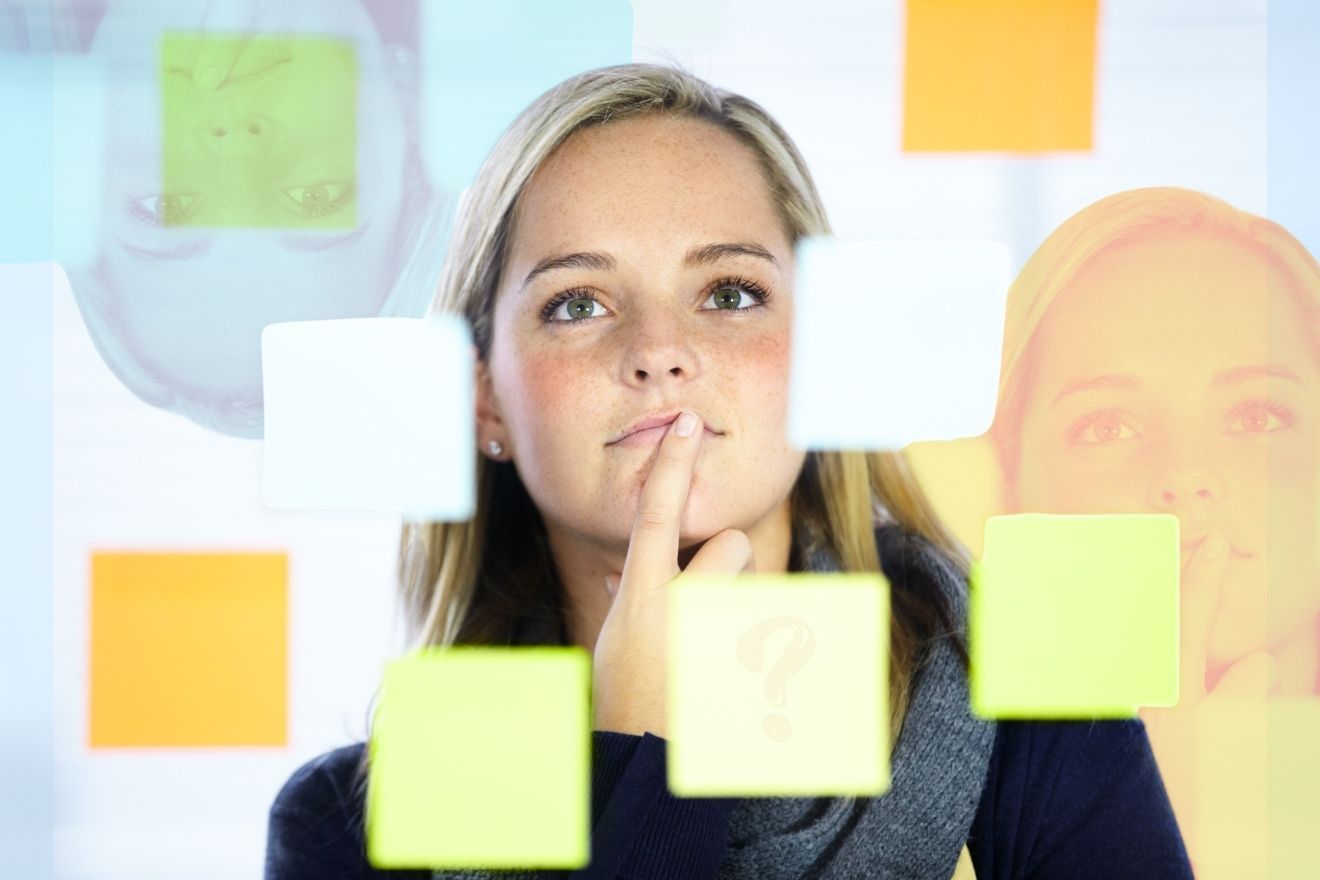 Are Psychedelics for Me? The image features a woman with blonde hair standing behind a transparent surface covered in colorful sticky notes, including shades of yellow, orange, green, and blue. She appears thoughtful, with her finger resting on her chin, suggesting she is reflecting or brainstorming. Her face is in focus, with a soft light illuminating her features. The composition includes blurred reflections of her face in the glass, adding depth and a dynamic visual element. The background is neutral, ensuring the sticky notes and her expression remain the focal points.