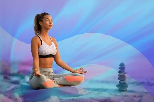 Spiritual Health Practitioners. The image depicts a woman in a meditative pose, sitting cross-legged on a yoga mat with her eyes closed and hands resting on her knees in a mudra position. She is wearing a sports bra and leggings. The background features a soft, dreamy gradient of blue, purple, and pink hues, creating a calming atmosphere. There is a stack of balanced stones to her right, adding a zen-like element to the composition. The scene conveys tranquility and mindfulness, with a focus on relaxation and meditation.