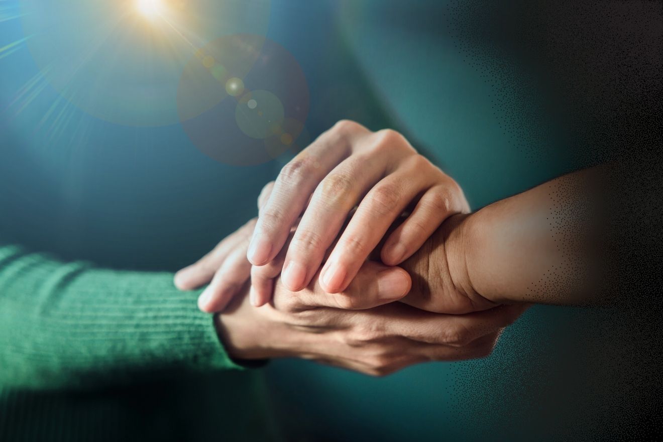 Risk of suicide. The image shows two clasped hands, radiating warmth and comfort. One hand, in a soft green sweater, rests on top of the other, which has a darker, natural tone. The deep blue-green background creates a peaceful feeling, while a soft light behind the hands adds a halo effect, symbolizing hope and protection. The overall mood is one of comfort, security, and deep connection, with a sense of optimism.