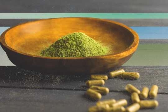 Kratom addiction. A wooden bowl filled with green powder beside scattered green capsules on a dark wooden surface, set against a striped background of muted blue, green, and gray tones.