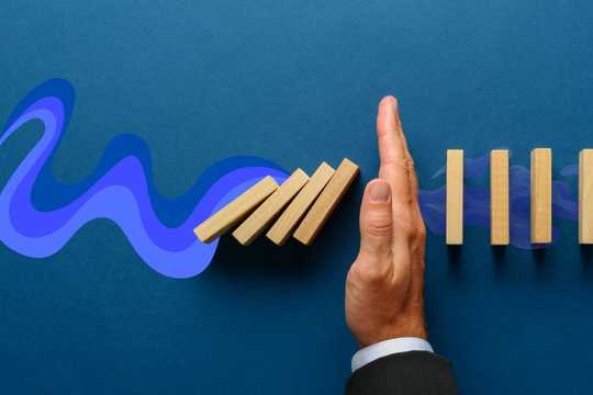 Integration Process. The image features a hand in a business suit stopping a row of falling wooden dominoes mid-collapse, symbolizing intervention or the prevention of a chain reaction. The background is a solid deep blue, with abstract wave-like shapes in varying shades of purple and blue flowing from left to right behind the dominoes. The composition creates a visual contrast between motion and stillness, with the hand serving as a clear focal point that interrupts the flow of energy and action.