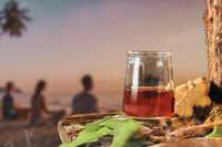 Ayahuasca Ceremony Etiquette. The image captures a serene sunset scene. In the foreground, a glass jar holds dark red ayahuasca brew, accompanied by pieces of ayahuasca bark and vibrant green leaves on a rustic wooden surface. In the background, three blurred figures sit on the beach, possibly meditating or conversing peacefully. The horizon features a soft, pastel sky with sunset hues of orange, pink, and purple, while a palm tree leans in from the corner, hinting at a tropical setting. This composition evokes a sense of calm and connection with nature, emphasizing relaxation and introspection.