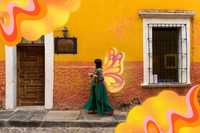Cultural Identity. Image of a person walking on a sidewalk next to a colorful warm yellow and orange building, which has rich wooden details. The person is wearing a long flowing deep green dress with a denim jacket over it, red shoes, and is carrying both a small dog and a take-away coffee. Another small dog is walking behind them on a leash. There are warm-coloured psychedelic pattern graphics in the top-left and bottom-right corners of the image, as well as butterfly wings which match from the back of the person.
