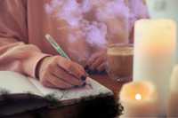 Shadow Work Journal Prompts. Image is of a person writing in a journal - it's a close up of their hands writing and the notebook. There are candles on the right, and a cup of a warm beverage too. Seeping from the pages of the notebook are black smokey cloud patterns. Rising up above the page are lilac smokey cloud patterns.