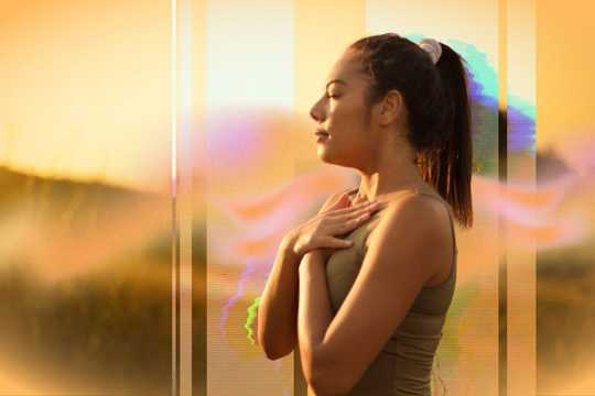 Holotropic Breathwork and Psychedelic Therapy. The image shows a woman standing outdoors at sunset, with her eyes closed and hands resting on her chest in a gesture of calm or meditation. Warm golden tones dominate the composition, with overlays of soft pinks, purples, and greens adding a surreal, dreamy quality. Vertical light streaks and subtle digital effects enhance the ethereal, introspective atmosphere of the scene.