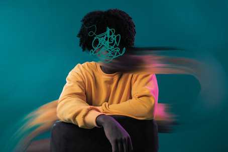Anxiety disorders. A teal background with a person sitting with their arms overlapping each other over their knees. They are wearing dark pants and a peachy-yellow long-sleeve top. They have big, dark curly hair. Their face is obscured by a mess of a squiggly line also in teal, and part of the image of them is blurred as if it was a painting that was disrupted by some horizontal strokes from a paintbrush.