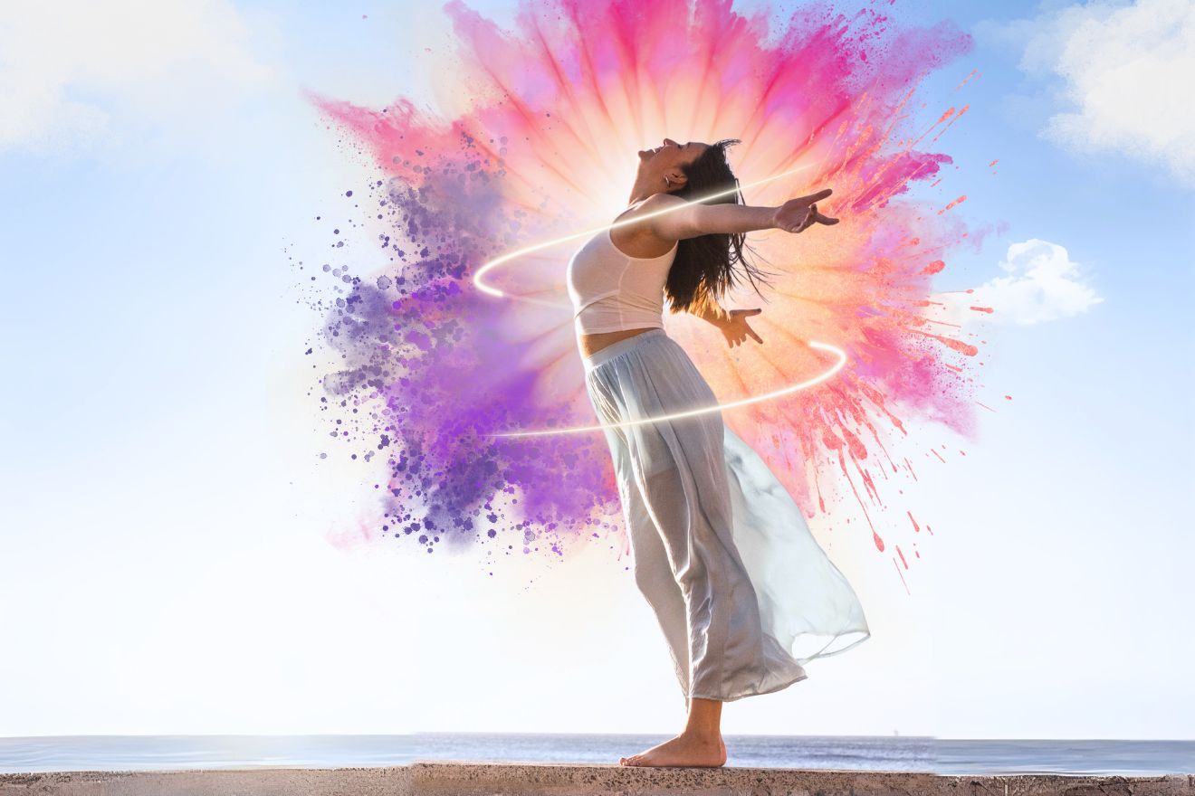 Personal Empowerment. Side-view photo of a female-presenting person with long hair standing on a beach with their arms thrown open wide in the air and their head raised. There is a graphic of pink and purple color explosions behind them and a glowing swirl circling them.