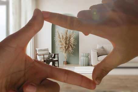 Setting in psychedelic. A pair of hands forms a rectangular frame in the foreground, visually “composing” a calm, minimalist interior scene behind them with soft beige and sage-green tones, natural light, and simple furniture arranged symmetrically for a serene, intentional feel.