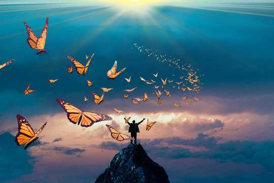 Concept of spiritual bypassing. Photograph of a person standing on the summit of a peak with their hands in the air. There is a soft pastel sunrise background behind them and butterflies all around them flying up into the sky.