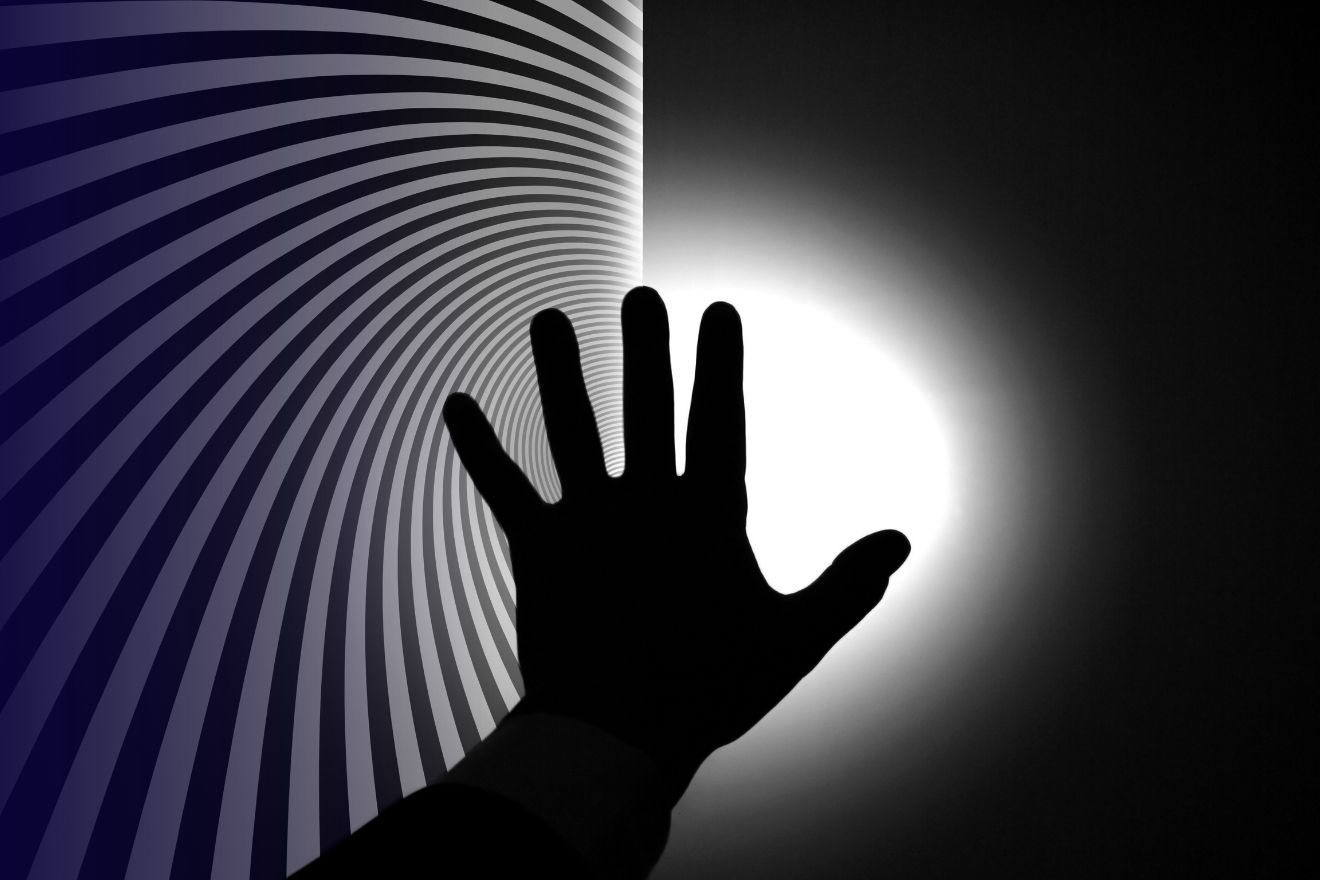 Near-Death and Psychedelic Experiences. The image features a silhouetted hand reaching towards a bright white light at the center-right, creating a strong contrast against a dark background. The left side of the image has a hypnotic spiral pattern of alternating dark and light grey lines that converge towards the light, drawing the eye inward. The hand is in the foreground, with fingers spread, and its dark silhouette is sharply outlined against the glowing white light, giving the scene a surreal and dramatic quality. The color scheme is monochromatic, dominated by black, grey, and white tones.