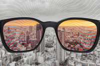 Psychedelics and Color Blindness. Image is of a city scape in greyscale, with a pair of glasses held up, where the photographer would be looking through the glasses at the city. Through the glasses, the city is in color, with a warm color palette.
