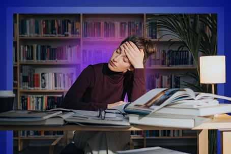 Burnout. A person sits at a desk in a library surrounded by open books, resting their head on one hand, with warm lamp light and purple-blue tones framing the scene.