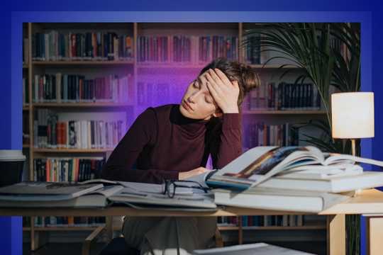 Burnout. A person sits at a desk in a library surrounded by open books, resting their head on one hand, with warm lamp light and purple-blue tones framing the scene.