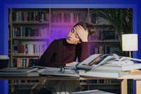 Burnout. A person sits at a desk in a library surrounded by open books, resting their head on one hand, with warm lamp light and purple-blue tones framing the scene.