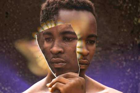 Self-Consent. The image shows a man holding a broken mirror shard that reflects his own face, creating a fragmented and introspective visual effect. The background is dark and textured with soft golden and purple light flares radiating from behind the mirror, giving the composition a surreal, dreamlike quality. The warm brown skin tones contrast against the cool, ethereal lighting, emphasizing themes of reflection, identity, and self-perception.